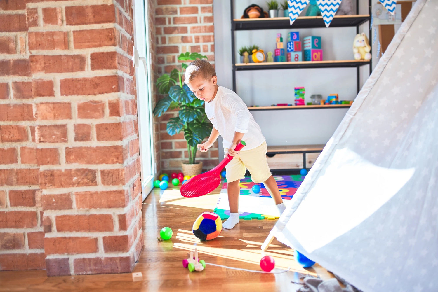 Child playing with colorful toys in a room with a brick wall and shelves.