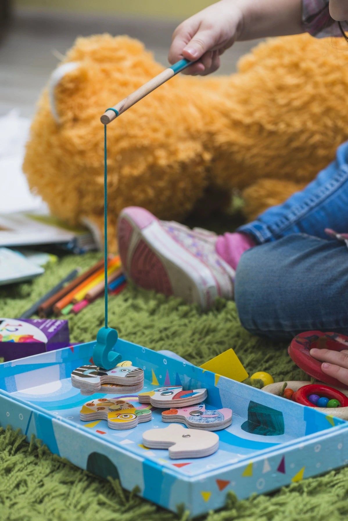 Child playing with a fishing game on the grass, surrounded by toys and a teddy bear.
