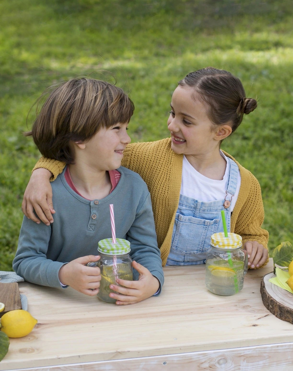 Two children sitting at a wooden table outdoors, enjoying drinks with straws.